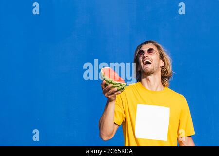 Attraktiver junger Mann mit langen blonden Haaren, die eine halbe Wassermelone in der Hand lächeln und sich auf einem schlichten blauen Hintergrund amüsieren. Sommer, Sonne, Stockfoto