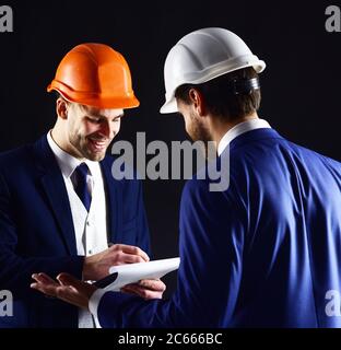 Ingenieure mit lächelnden Gesichtern diskutieren über das Projekt. Chef im Helm gibt Anweisungen an den Mitarbeiter. Geschäftsmann und glücklicher Architekt mit Architekturprojekt. Bau, Architektur, Arbeit, Arbeitskonzept. Stockfoto