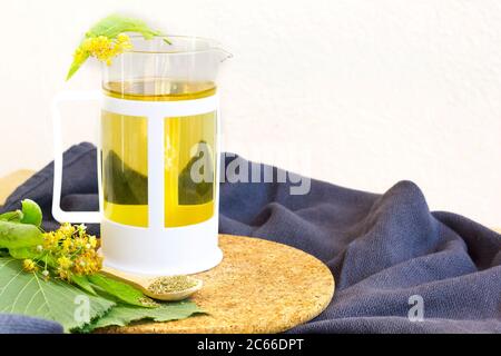 Teekanne aus Glas mit Lindentee und Blumen auf dem Tisch im Garten. Heller Hintergrund. Platz für Text. Bio-Tee mit frischen Lindenblüten. Kräutertee Stockfoto