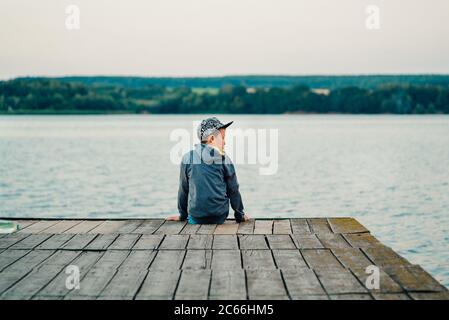 Der kleine Junge sitzt auf der Brücke in der Nähe des Sees. Er ist von einer wunderbaren Aussicht umgeben Stockfoto