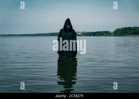 Ein Mann in einer schwarzen Robe mit Kapuze steht tagsüber im Fluss. Er schaut auf mysteriöse Weise auf das Wasser Stockfoto