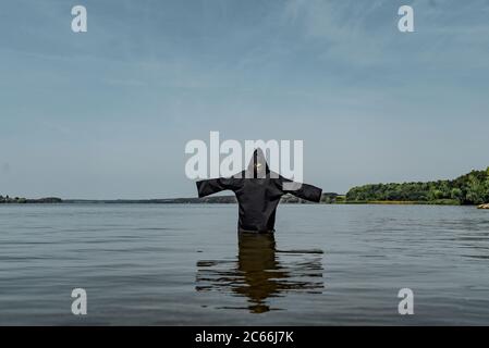 Ein Mann in einer schwarzen Robe mit ausgestreckten Händen steht bei warmem Wetter mitten im See. Sein Blick ist auf das Wasser gerichtet Stockfoto