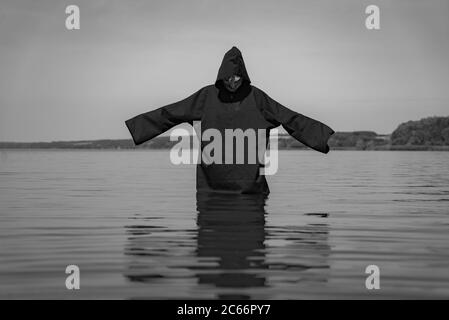 Ein Geist in einem schwarzen Mantel steht im See mit Händen auf dem Hintergrund der Natur angehoben. Das Phantom ist in einem Schwarz-Weiß-Foto dargestellt. Stockfoto