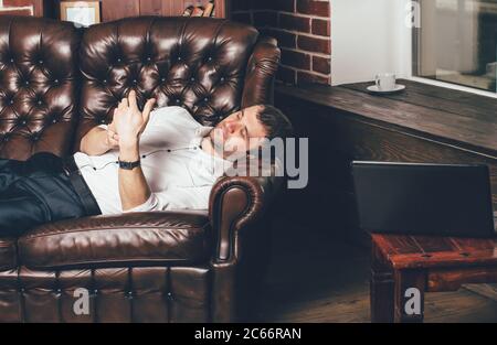 Der Geschäftsmann ruht auf einem Ledersofa in der Nähe eines Laptops und eine Tasse Tee im Zimmer. Der Mann hält das Telefon in der Hand. Stockfoto