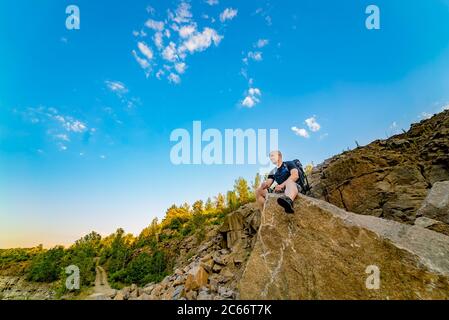 Ein Reisender mit Rucksack sitzt auf einem großen Stein mit einem scharfen Ende auf dem Hintergrund von Felsen und blauen Himmel im Sommer Stockfoto