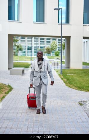 Afroamerikanisch in einem weißen Anzug und einem roten Koffer. Geschäftsmann. Geschäftsreisende. Reisender Mann. Stockfoto