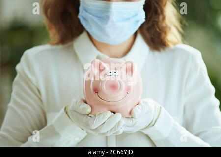 Leben während einer Coronavirus-Pandemie. Nahaufnahme der Frau in weißer Bluse mit medizinischer Maske, Sparschwein und Handschuhen. Stockfoto