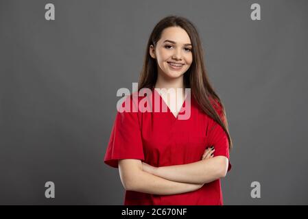 Porträt einer selbstbewussten jungen und lächelnden Krankenschwester mit roten Scrubs auf grauem Hintergrund Stockfoto