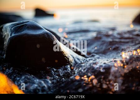 Nahaufnahme von Meereswellen, die auf einer nassen Küste brechen Steine Sommer Sonnenuntergang, Wasserspritzer, Sonnenstrahlen, Reflexionen auf Wassertropfen. Stockfoto