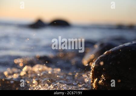 Nahaufnahme von Meereswellen, die auf einer nassen Küste brechen Steine Sommer Sonnenuntergang, Wasserspritzer, Sonnenstrahlen, Reflexionen auf Wassertropfen. Stockfoto