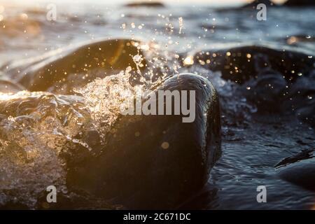 Nahaufnahme von Meereswellen, die auf einer nassen Küste brechen Steine Sommer Sonnenuntergang, Wasserspritzer, Sonnenstrahlen, Reflexionen auf Wassertropfen. Stockfoto