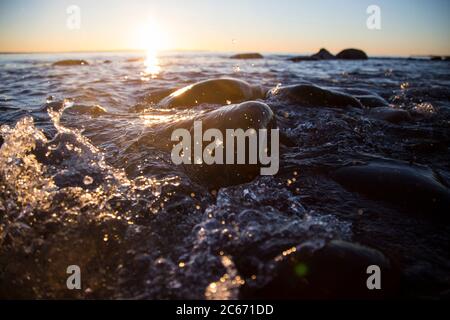 Nahaufnahme von Meereswellen, die auf einer nassen Küste brechen Steine Sommer Sonnenuntergang, Wasserspritzer, Sonnenstrahlen, Reflexionen auf Wassertropfen. Stockfoto