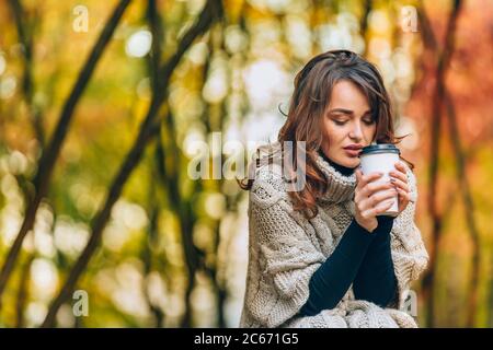 Schöne Frau in einem Strickpullover hält ein Glas heißen Kaffee im Park im Herbst. Ein Mädchen wärmt sich mit einem heißen Getränk Stockfoto