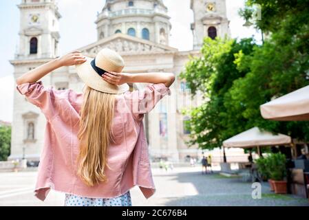 Rückansicht einer langhaarigen Frau mit Strohhut, während sie mit ausgestreckten Armen vor der St.-Stephans-Basilika in Budapest stand. Stockfoto