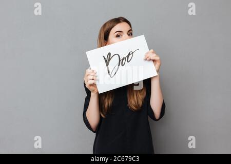 Portrait von hübschen Mädchen stehen und ihr Gesicht zu bedecken, während Postkarte auf grauem Hintergrund halten Stockfoto