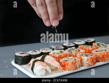 Nahaufnahme des Prozesses der Zubereitung von Sushi-Rollen. Der Koch dekoriert köstliche frische Brötchen mit Sesam auf dem Holztisch. Dunkler Hintergrund Stockfoto