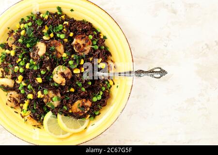 Sommerliches Speisekonzept. Kalter schwarzer Reissalat mit Mais, Erbsen, Garnelen und Zitrone auf gelbem Teller. Gesunde Ernährung. Draufsicht. Stockfoto