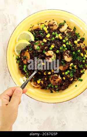 Sommerliches Speisekonzept. Kalter schwarzer Reissalat mit Mais, Erbsen, Garnelen und Zitrone auf gelbem Teller. Gesunde Ernährung. Draufsicht. Stockfoto