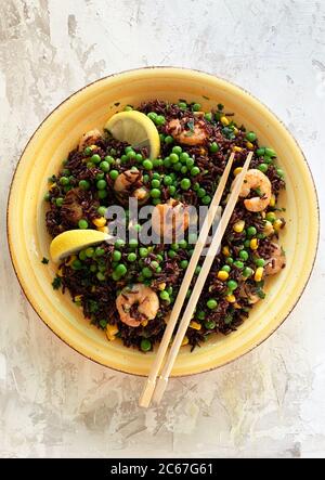 Sommerliches Speisekonzept. Kalter schwarzer Reissalat mit Mais, Erbsen, Garnelen und Zitrone auf gelbem Teller. Gesunde Ernährung. Draufsicht. Stockfoto