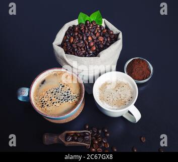 Heiße Kaffeetassen und Kaffeebohnen in Leinenbeutel und gemahlenes Pulver auf schwarzem Küchentisch Hintergrund. Stockfoto