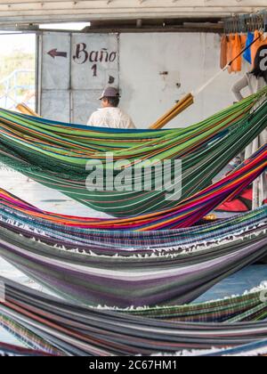 Amazonas-Fluss, Peru - 12. Dez 2017: Der Mann in der Spitzenmütze sitzt hinter schönen, bunten Hängematten auf dem Frachtschiff. Amazonien, Wanderweg von Santa R Stockfoto