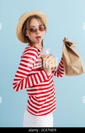 Junge hübsche Mädchen mit zwei Zöpfen in Strohhut und rote Sonnenbrille hält Orangensaft und Papiertüte mit Lebensmitteln in den Händen über blauen Hintergrund isoliert Stockfoto