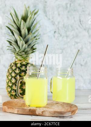 Ananassaft in Maurer-Glas-Gläsern mit Metallstrohhalmen und Ananasfrucht auf Holzbrett auf blauem Marmorhintergrund. Erfrischungscocktail im Sommer. Stockfoto