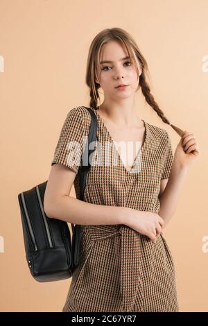 Junge schöne Mädchen mit zwei Zöpfen in Tweed Jumpsuit mit schwarzem Rucksack auf der Schulter traumhaft in der Kamera über beige Hintergrund isoliert suchen Stockfoto