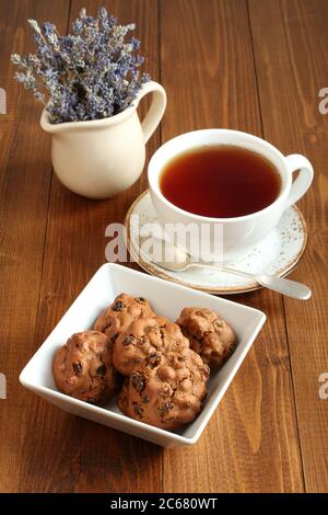 Schokoladenkekse mit Walnüssen und Rosinen in einer quadratischen Schüssel und eine Tasse schwarzen Tee auf einem Holztisch. Nahaufnahme Stockfoto
