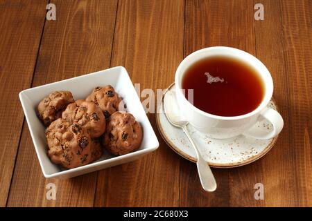 Schokoladenkekse mit Walnüssen und Rosinen in einer quadratischen Schüssel und eine Tasse schwarzen Tee auf einem Holztisch. Nahaufnahme Stockfoto