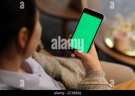 Über der Schulter einer jungen Frau hält ein grünes Bildschirm Smartphone im Wohnzimmer in der Nacht. Weichzeichnen Hintergrund Stockfoto