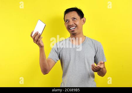 Lustige junge asiatische Mann hält Smartphone, isoliert auf gelbem Hintergrund Stockfoto