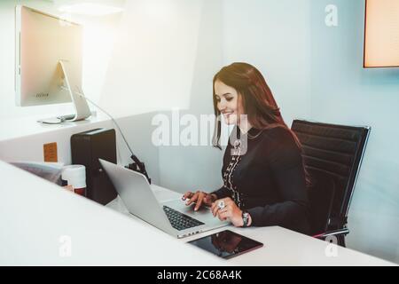 Die freundliche, schöne Rezeptionistin mit langen Haaren lächelt, während sie auf einem Sessel, umgeben von Gadgets, sitzt und drinnen einen Laptop benutzt Stockfoto