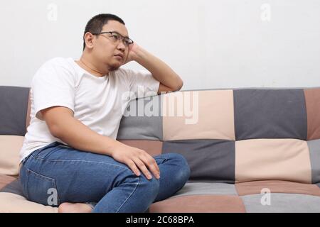 Unglücklicher asiatischer Mann, der zu Hause im Wohnzimmer auf seinem Sofa sitzt und denkt, trauriger, besorgter Ausdruck Stockfoto