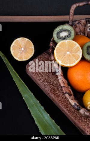 Ein gewebter Korb voller frischer Orangen, Zitronen und grüner Kiwis in der Nähe eines Aloe Vera Blattes auf schwarzem Hintergrund. Stockfoto