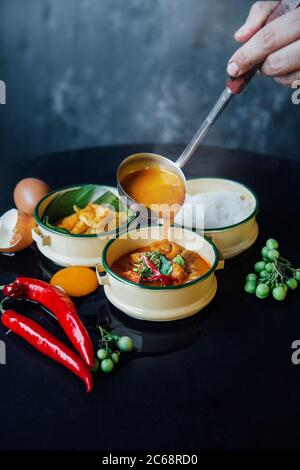 Schöpfkelle gießen Rote Currypaste Servieren in Tiffin-Box mit rohen Zutaten auf schwarzem Tisch Stockfoto