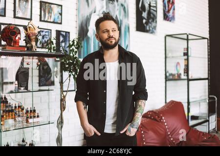 Junge Tätowierer in schwarzem Hemd nachdenklich beiseite schauen Zeit in Tattoo-Studio verbringen Stockfoto