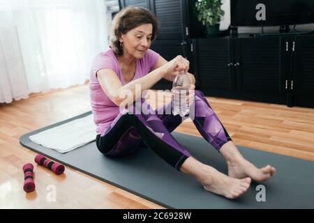 Ältere Frau wird nach dem Training zu Hause Wasser trinken. Müde ältere Frau in lila Sportkleidung sitzt auf Fitness-Matte Öffnung Flasche mit Wasser Stockfoto