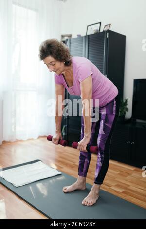 Aktive ältere Frau macht Training zu Hause. Ganzkörperaufnahme der fokussierten 70 Jahre alte Frau in lila Sportbekleidung Training ihren Rücken halten Hanteln bo Stockfoto