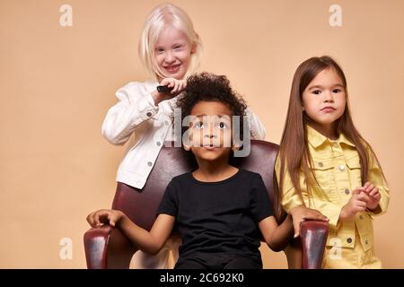 Portrait von freundlichen, vielfältigen Kindern spielen Friseure. Zwei Mädchen kämmen Jungen lockiges Haar, glücklich lächelnde Kinder mit ungewöhnlich vielfältigen Aussehen. natur Stockfoto
