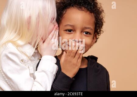 Portrait von überrascht afro Junge und positive Albino Mädchen mit Geheimnis. Freundliches Mädchen erzählen Geschichte in Jungen Ohren, Junge stehen in Schock schließenden Mund Stockfoto