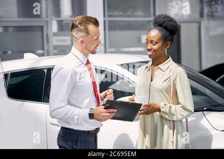 Junge schöne attraktive afrikanische Dame trinken eine Tasse Tee und aufmerksam zuhören, professionelle Vertriebsmitarbeiter, spricht Mann über Funktionen des Autos Stockfoto