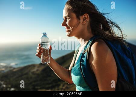 Frau auf der Klippe und Trinkwasser. Bergsteigerin mit einer Wasserflasche in der Hand und Blick auf die schöne Aussicht. Stockfoto