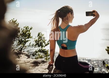 Rückansicht einer fittenden jungen Frau, die auf der Klippe sitzt und Selfie nimmt. Frau in Sportbekleidung macht ein Selbstporträt mit Handy auf Berggipfel. Stockfoto