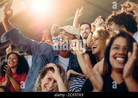 Gruppe von Fans beobachten Fußballspiel und machen Selbstporträt mit Smartphone im Stadion. Multiethnische Menschen genießen ein Spiel im Stadion Stockfoto