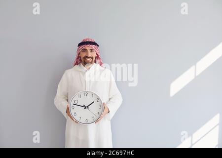 Schöner arabischer Mann, der eine Uhr in den Händen hält Ein grauer Hintergrund Stockfoto