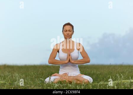 Frau Yoga in der Natur. Ein Mädchen in weißen Kleidern praktiziert Meditation, während es in einer Lotusposition auf dem Gras gegen den Himmel sitzt. Stockfoto