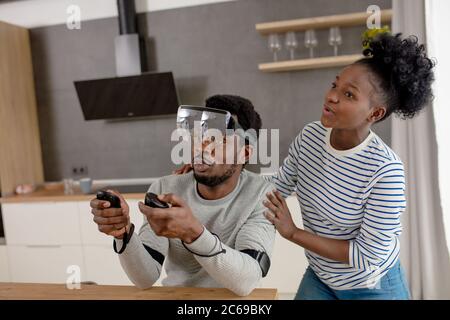 Afrikanischer mann Vornehmen der Einstellungen der virtuellen Realität Konsole mit Controller, während seine Frau ihm die Vorbereitung am Morgen Kaffee in der modernen Küche Stockfoto