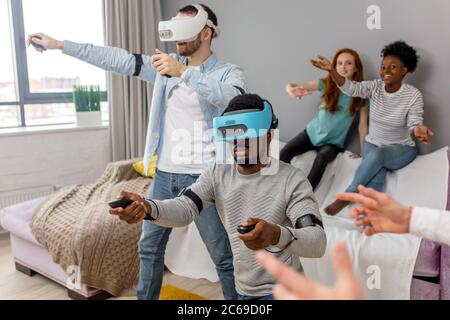 Multiethnischen diverse Freunde genießen die VR-Technologie, zwei Kerle tragen VR-Headset und spielen, während die afrikanischen und kaukasische rothaarige Mädchen schauen Sie an und Stockfoto