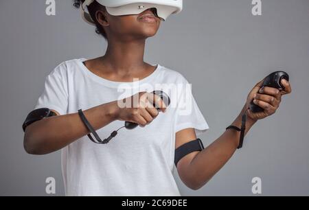 Junge afrikanische Studentin in weißem T-Shirt mit Virtual-Reality-Brille Headset spielen, Hände in Schießweise halten, als ob sie ein Gewehr hält, p Stockfoto